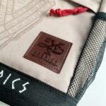 Close-up of an embossed brown leather Nordics logo patch on a beige canvas backpack with a red zipper pull.